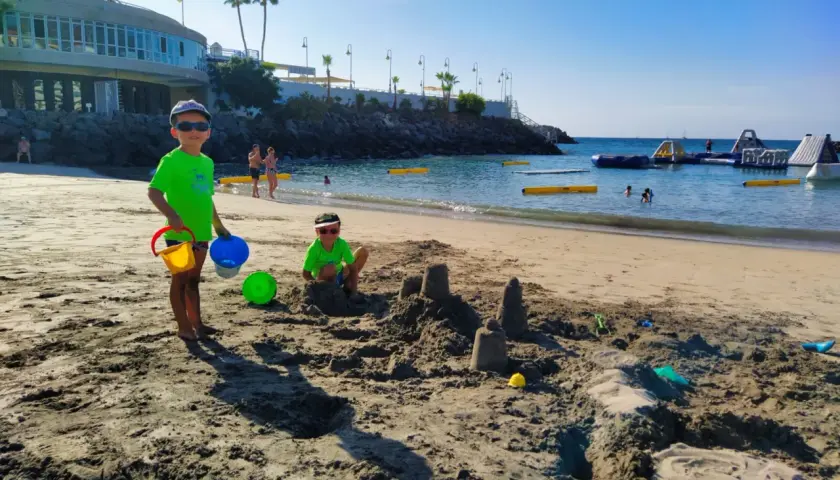Radosne **teneryfa z dziećmi** na plaży. Dwóch chłopców buduje zamki z piasku, a w tle widać wodne atrakcje.