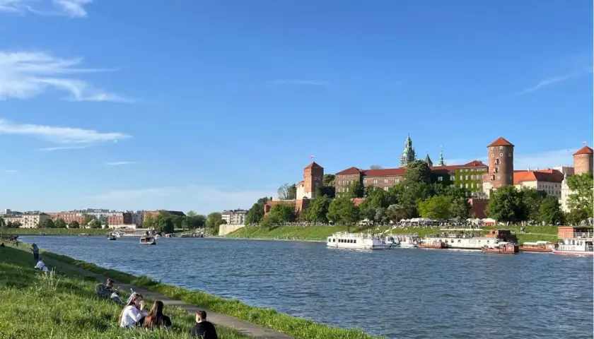Słoneczny dzień nad Wisłą, ludzie odpoczywają na trawie, a w tle majaczy Wawel – symbol starego miasta Krakowa.