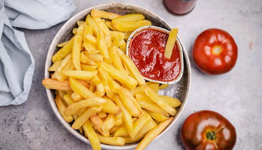 Knusprige Pommes Frites mit einem Schälchen veganem Ketchup. Zwei Tomaten und eine Flasche Ketchup liegen daneben.