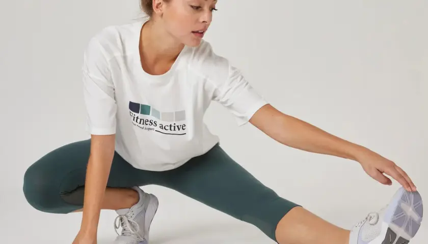 Kobieta w białej koszulce z napisem "Fitness active" wykonuje rozciąganie po treningu siłowym.