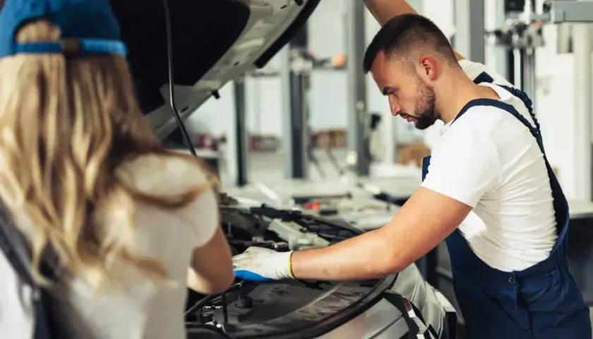 Mechanik z rękawiczkami bada silnik, szukając przyczyny usterki systemu zarządzania silnikiem. Obok stoi kobieta w czapce.