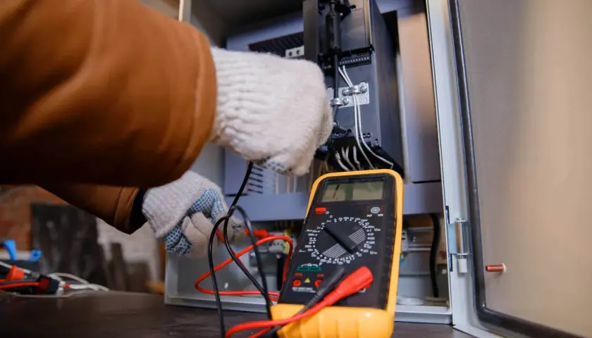 Elektryk w rękawiczkach sprawdza połączenia w szafie sterowniczej za pomocą żółtego multimetru. Pokazuje, jak używać multimetru do pomiarów.