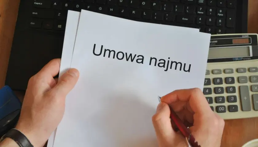 Ręka pisze długopisem umowę najmu. Obok leży kalkulator i klawiatura, gotowe do pracy dla wynajmującego.