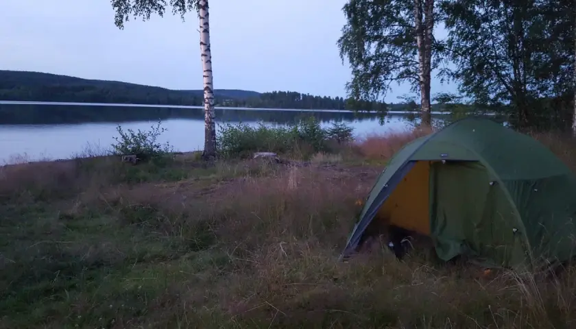 En grön campingtält står vid en sjö, med en skogklädd kulle i bakgrunden. En perfekt plats för äventyr längs 7 torpsleden.