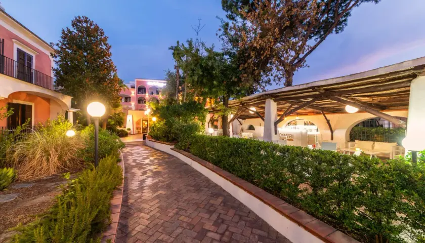 Sentiero in mattoni che porta a un bar all'aperto illuminato, con edifici color pastello sullo sfondo. Un'atmosfera da sogno al Valentino Ischia.
