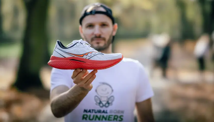 Mężczyzna w koszulce "Natural Born Runners" prezentuje biało-czerwony but biegowy Saucony. Saucony to znana firma z tradycjami.
