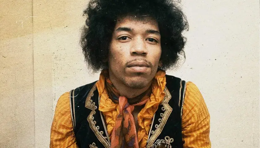 Jimi Hendrix w pomarańczowej koszuli i kamizelce, z charakterystyczną fryzurą. W tle subtelna, lekko zatarte tło, jakby w **purple haze**.