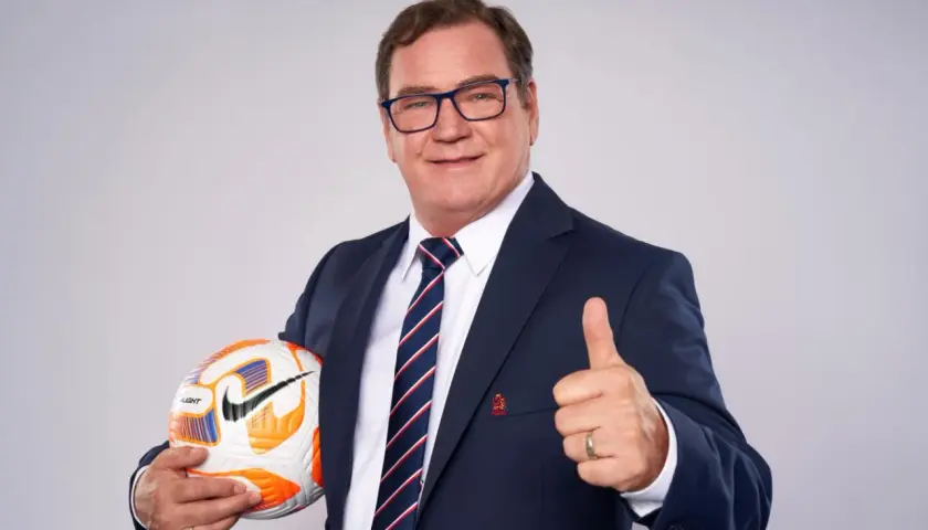 Uśmiechnięty mężczyzna w garniturze, z piłką w ręku i kciukiem w górze. Wygląda jak trener reprezentacji Polski, gotowy do akcji.