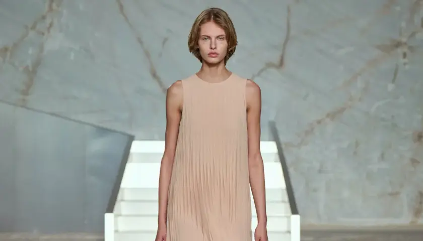 Modelo con vestido plisado color melocotón, en un escenario minimalista con escaleras blancas y fondo de mármol.