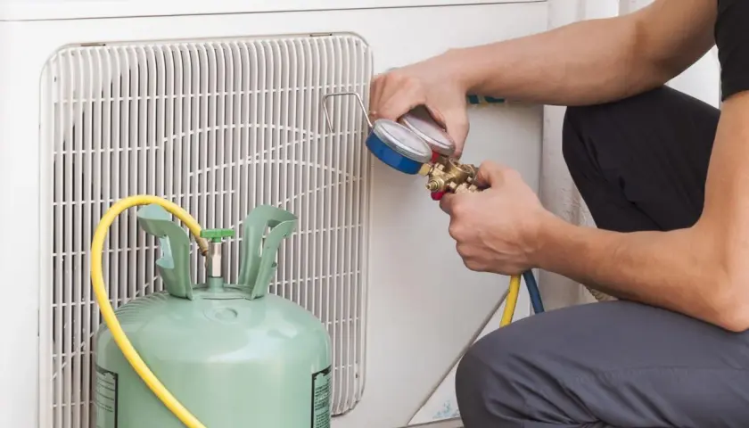 Técnico revisa manómetro para detectar fuga aire acondicionado. Cilindro de gas refrigerante y unidad exterior visibles.