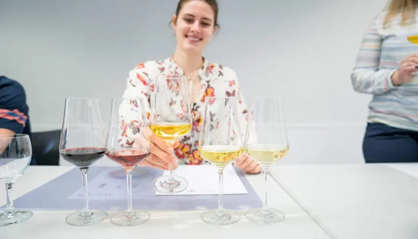 Eine Frau mit Blumenmuster-Bluse verkostet Wein, ein Schritt auf dem Weg, Sommelier zu werden. Mehrere Gläser mit Rot-, Rosé- und Weißwein stehen auf dem Tisch.