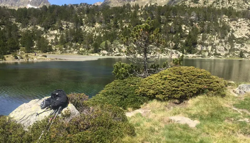 Mochila y bastones de senderismo junto a un llac de pessons, rodeado de pinos y montañas rocosas.