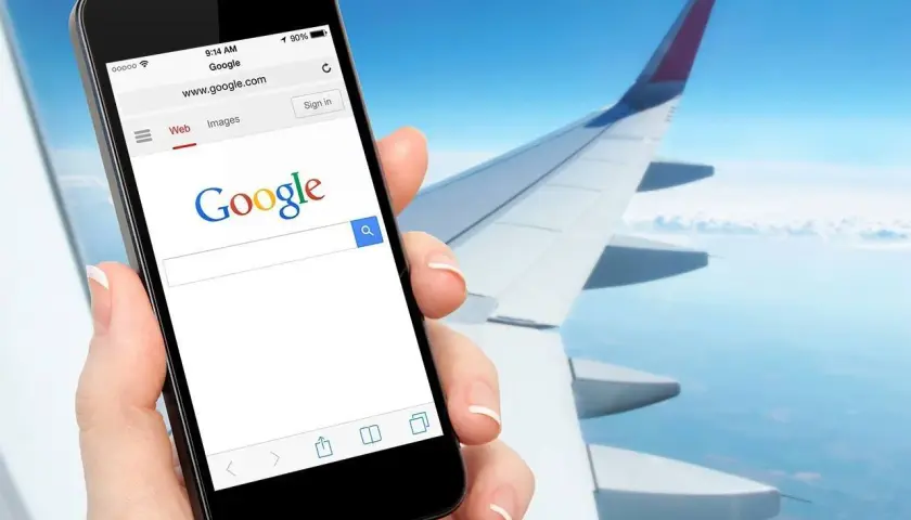 Dłoń trzyma smartfon z otwartą wyszukiwarką Google, na tle skrzydła samolotu i nieba. Czy w samolocie można mieć internet? Tak, można!