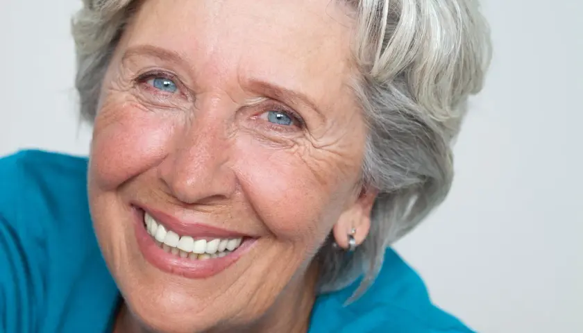 Donna anziana con capelli grigi e occhi azzurri, sorride mostrando le rughe intorno alla bocca, segno di una vita vissuta con gioia.