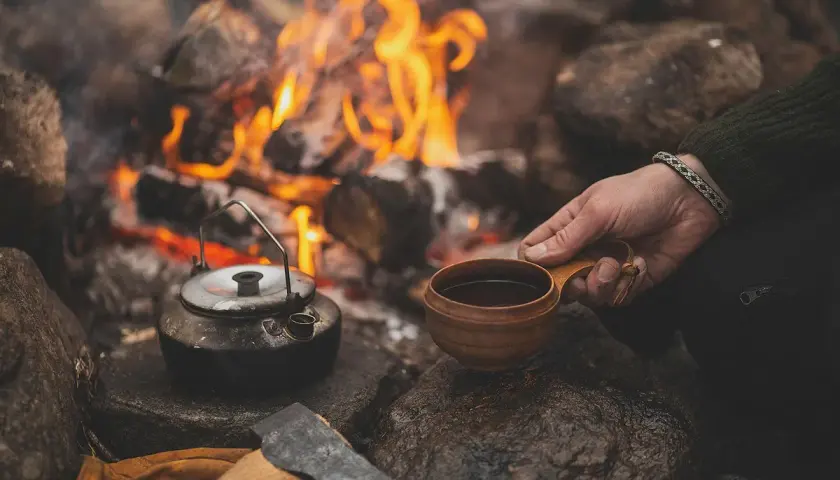 En hand håller en träkopp med kaffe vid en lägereld. En kaffepanna står bredvid, redo att koka kaffe på gammalt vis.