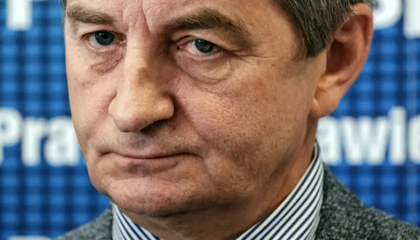 Mężczyzna w garniturze, w tle niebieska krata z białymi literami. Czy to on wyjaśnia, dlaczego posłowie PiS zostali oskarżeni o nadużywanie lotów rządowych?
