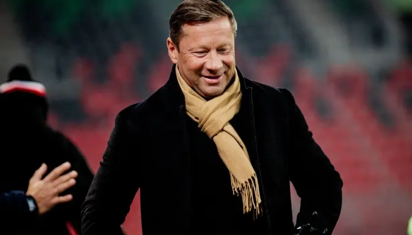 Uśmiechnięty Dariusz Banasik w czarnym płaszczu i beżowym szaliku, na tle trybun stadionu.