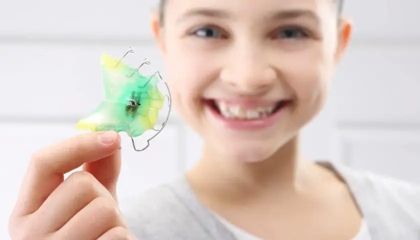 Niña sonriente muestra un retenedor dental verde y amarillo, listo para limpiar.