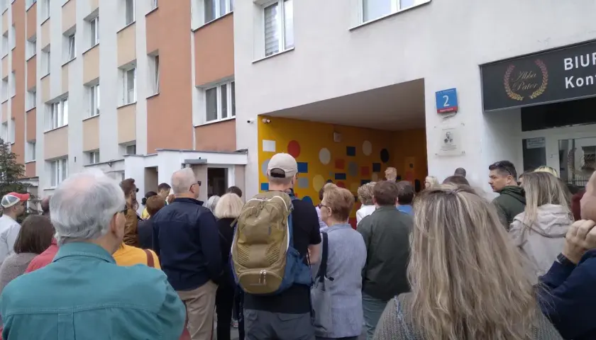 Grupa ludzi stoi przed blokiem mieszkalnym z kolorową ścianą i numerem 2.