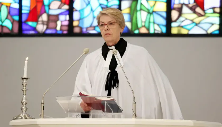 Kobieta w stroju duchownego, z białą narzutą i czarną kryzą, stoi przy pulpicie. To pastor, który przygotowuje się do wygłoszenia kazania.