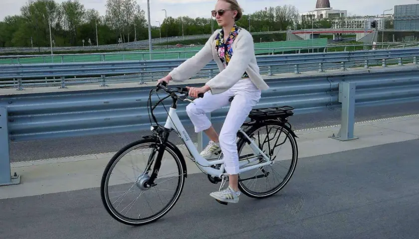 Kobieta w białych spodniach i sweterku jedzie na rowerze elektrycznym. W tle most i budynki.