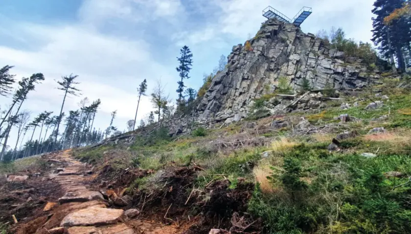 Kamienista ścieżka prowadzi ku skalistemu szczytowi z platformą widokową. W oddali widać las, a krajobraz przypomina ten z **Trójaka w Lądku Zdroju**.