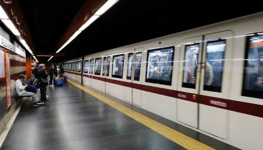 Des voyageurs attendent sur le quai du **metro a Rome**. Un train blanc et bordeaux arrive, reflétant les lumières du tunnel.