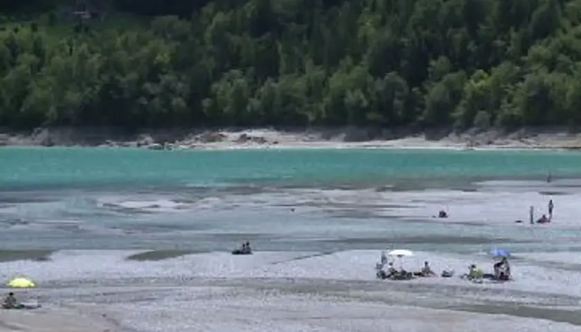Persone rilassate sulla spiaggia del lago di Barcis, con acque turchesi e boschi sullo sfondo.