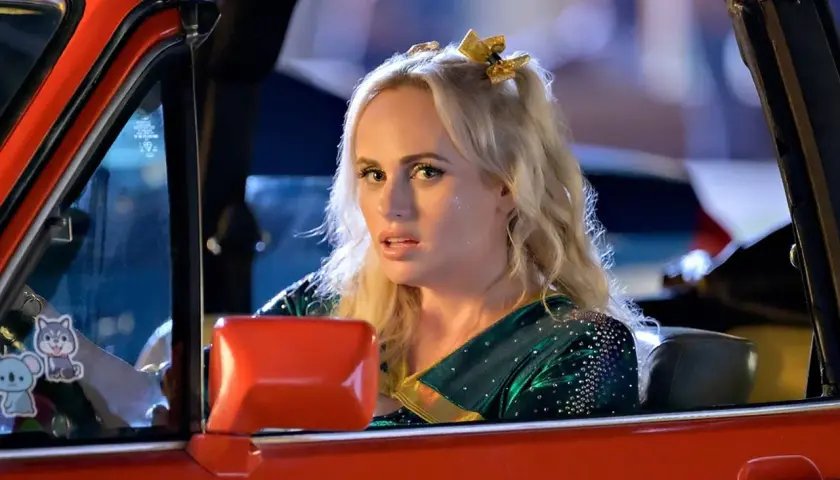Dziewczyna w stroju cheerleaderki, z błyszczącymi akcentami, jedzie czerwonym kabrioletem. To jej powrót do liceum po latach.