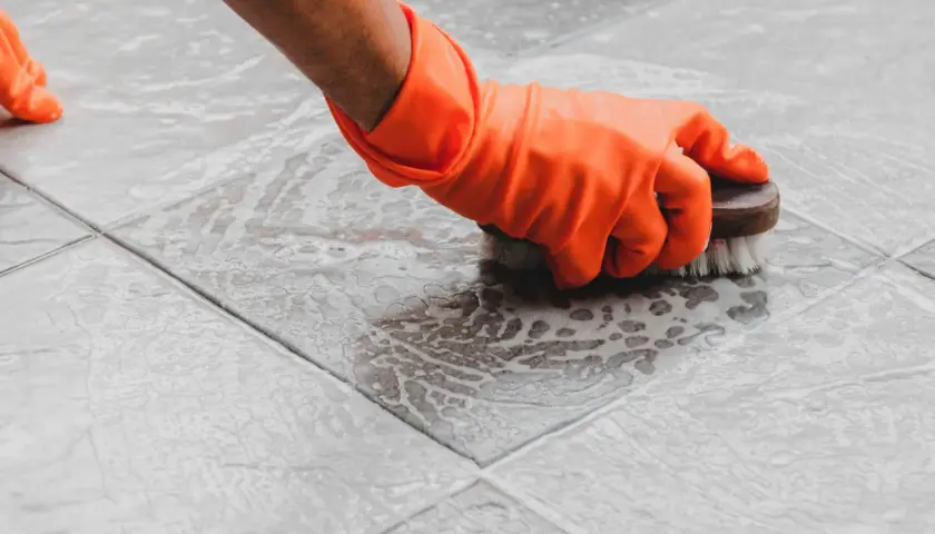 Mano con guanto arancione che lucida le piastrelle del pavimento con una spazzola, creando schiuma.