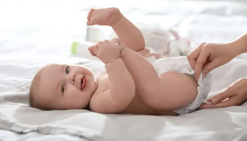 Uśmiechnięte dziecko leży na plecach, a ręka mamy zmienia mu pieluszkę, zapobiegając pieluszkowemu zapaleniu skóry.