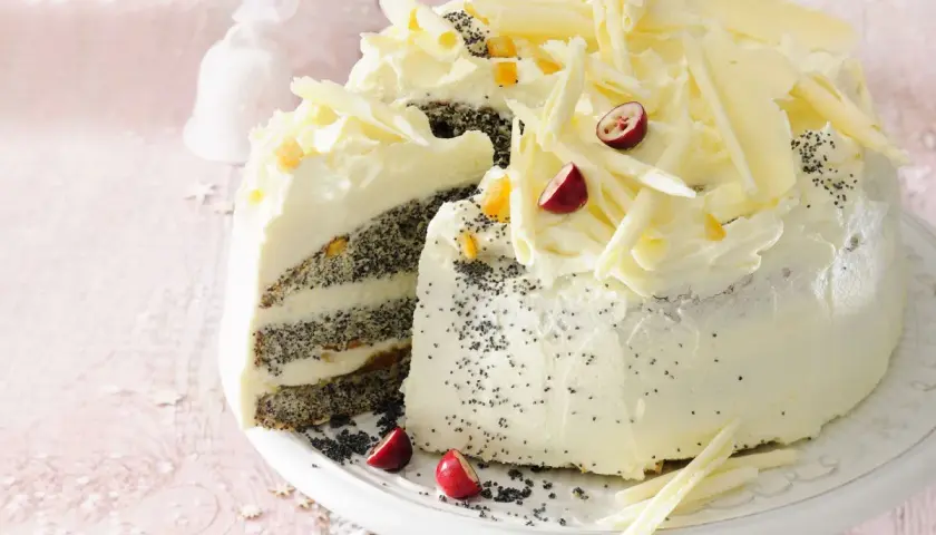 Pyszny tort makowy mojej mamy, udekorowany kremem, białymi czekoladowymi wiórkami, żurawiną i kandyzowaną skórką pomarańczową.