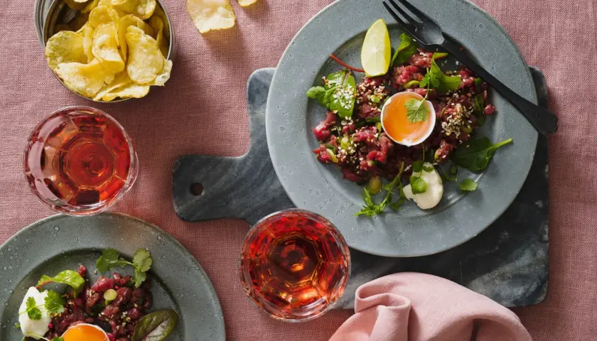 Två tallrikar med asiatisk råbiff, toppad med äggula och sesamfrön. Serveras med potatischips och rosévinsglas.