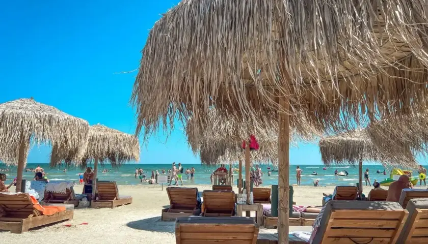 Słoneczne rumuńskie wakacje nad Morzem Czarnym. Leżaki i parasole z trawy na piaszczystej plaży, w tle ludzie kąpiący się w morzu.