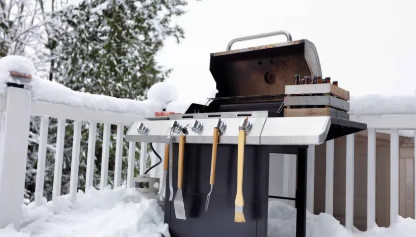 Un barbecue d'hiver prêt à l'emploi, avec des bouteilles de bière dans une caisse en bois et des ustensiles. La neige recouvre la terrasse et la balustrade.