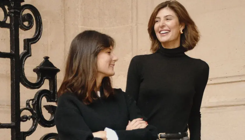 María de la Orden y su amiga ríen juntas frente a una reja ornamental.