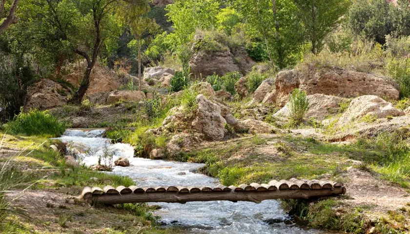 Un puente rústico cruza un arroyo en un paisaje rocoso y verde, evocando la serenidad de una cascada en Chelva.