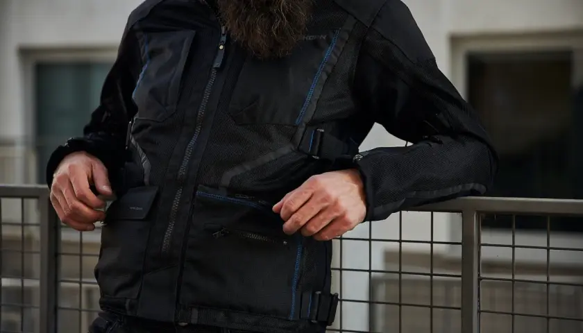 Mężczyzna w czarnej kurtce motocyklowej Rebelhorn Scandal II z niebieskimi akcentami, oparty o metalową barierkę.