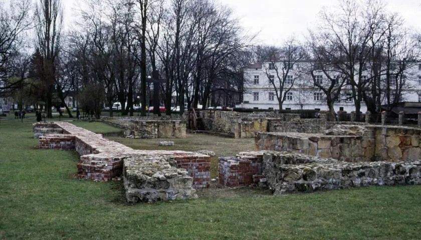Ruiny zamku w Wieluniu, kamienne i ceglane fundamenty na trawiastym terenie, w tle drzewa i biały budynek.