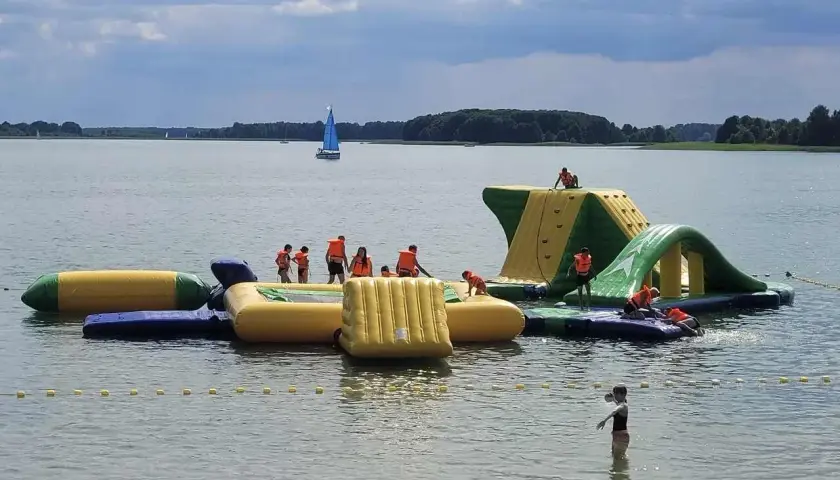 Dzieci w kamizelkach ratunkowych bawią się na dmuchanych zjeżdżalniach i torach przeszkód na jeziorze. Idealne miejsce, gdzie na Mazury z dziećmi!