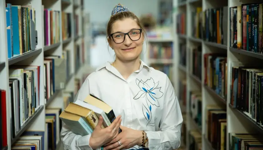 Bibliotekarka z okularami trzyma stos książek. W tle regały pełne literatury. Czy wakat co to znaczy?