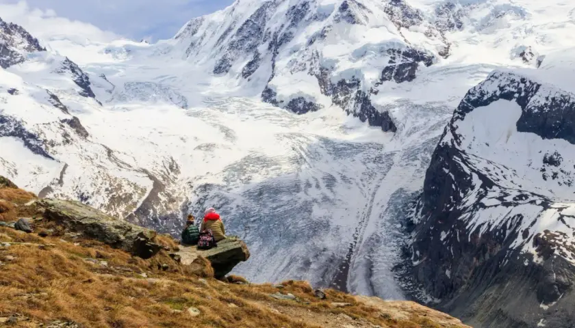 Two hikers rest on a rocky outcrop, gazing at the majestic, snow-covered peaks of the tour de Monte Rosa.