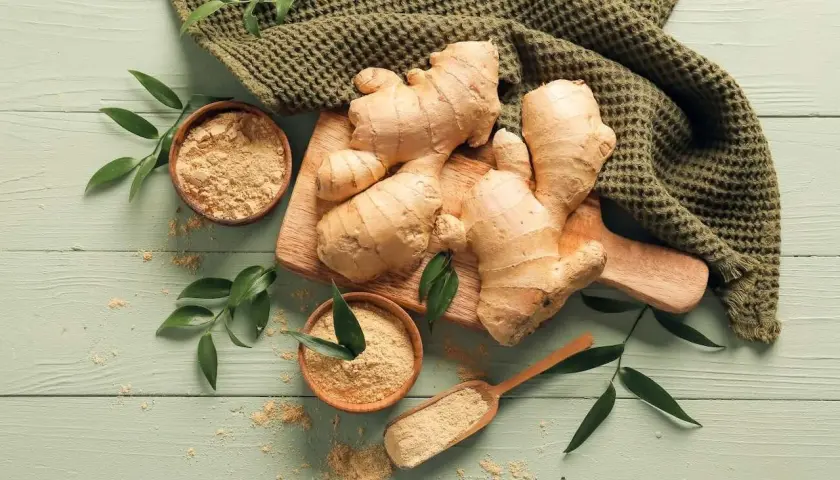 Imbir na co pomaga? Świeże korzenie i mielony imbir na drewnianym tle, otoczone zielonymi liśćmi.