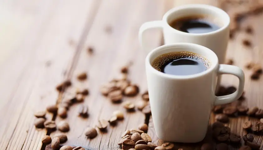 Dwa białe kubki z aromatycznym espresso na drewnianym blacie, otoczone ziarnami kawy. Zastanawiasz się, ile mg kofeiny ma espresso?