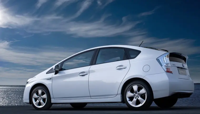 Biały Prius 3 zaparkowany nad wodą, pod błękitnym, chmurnym niebem.