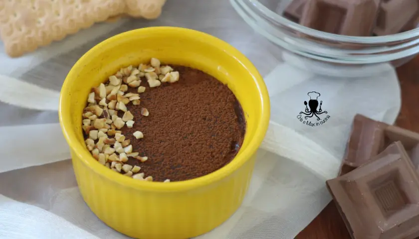 Budino al cioccolato in ciotola gialla, cosparso di cacao e nocciole tritate. Accanto, biscotti e cioccolato.