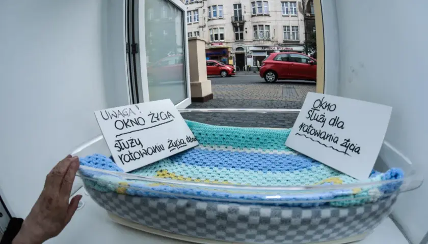 Okno życia z tabliczkami informacyjnymi w koszu, widok na ulicę z budynkami i samochodami.