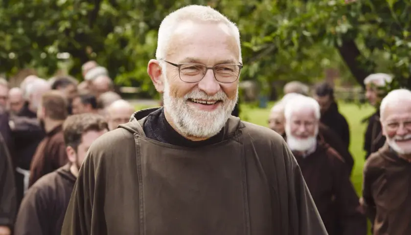 Bruder Paulus, ein älterer Mann mit Brille und Bart, lächelt in die Kamera. Hinter ihm stehen weitere Männer in braunen Kutten.