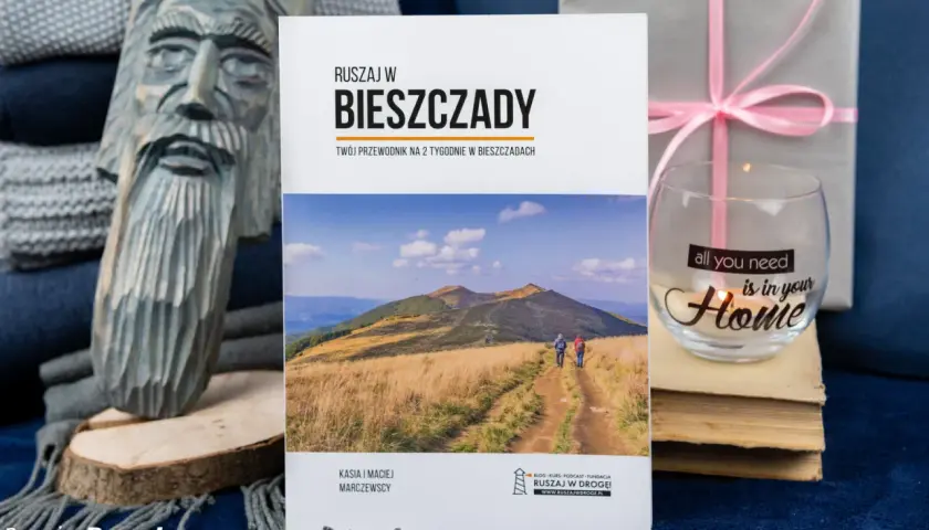 Przewodnik "Ruszaj w Bieszczady" z informacjami o Bieszczadzkim Parku Narodowym. Na zdjęciu także rzeźba, szklanka z napisem i książki.