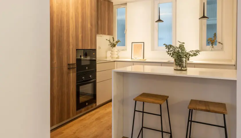 Cocina moderna con muebles de madera, encimera blanca y dos taburetes. La luz fría k ilumina el espacio, creando un ambiente acogedor.